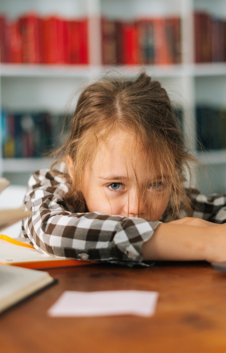 A frustrated and worn out girl rests her head on her arms at a cluttered desk with books and a laptop in a classroom.