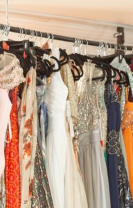 Colorful evening gowns hang on a boutique rack, featuring sequins, beadwork, sheer fabric, and flowing skirts.