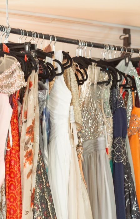 Colorful evening gowns hang on a boutique rack, featuring sequins, beadwork, sheer fabric, and flowing skirts.