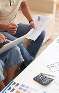A man and woman sitting on a couch with a laptop. They are sitting in front of building blueprints for their home.