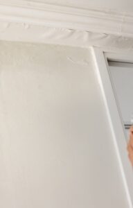 A woman wearing glasses covers her mouth while she looks at water damage and cracks on a white ceiling.