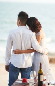 A couple standing with arms around each other beside a beach table with plates, wine, and ocean waves behind them.
