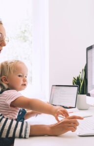 A mom wearing glasses sits with a child and works at home next to a white desk beside a large monitor and tablet.