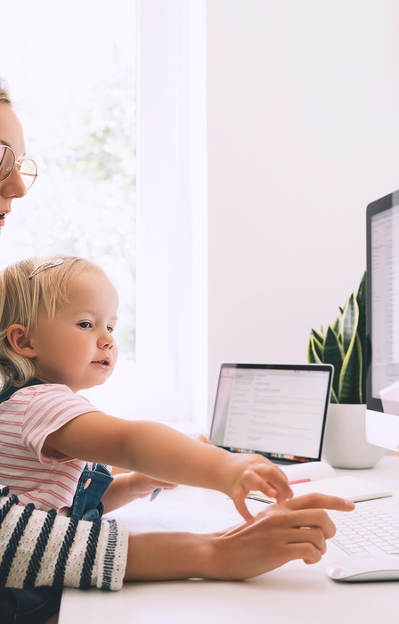 A mom wearing glasses sits with a child and works at home next to a white desk beside a large monitor and tablet.