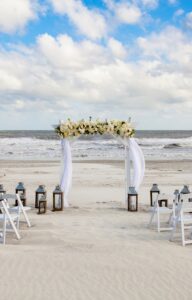 A wedding setup on a beach with clouds in the sky. There is an altar, two rows of chairs, and several lanterns.
