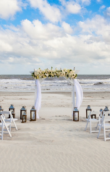 A wedding setup on a beach with clouds in the sky. There is an altar, two rows of chairs, and several lanterns.