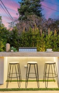 A small, outdoor bar in a home's backyard. The modern bar is made completely of stone and features three stools.