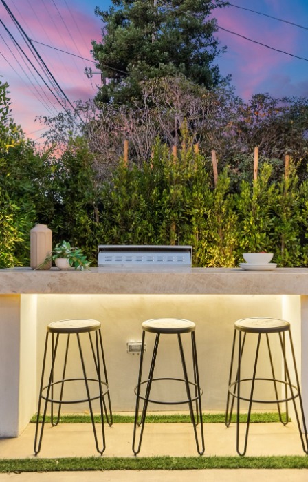 A small, outdoor bar in a home's backyard. The modern bar is made completely of stone and features three stools.