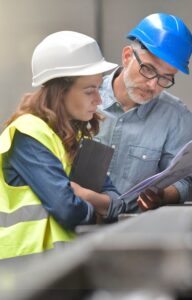 Two employees wearing hard hats in a brightly lit factory. Both employees look at a large paper document.