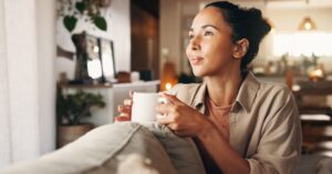A woman sitting on a couch and holding a white mug while looking toward a bright window in a cozy home.