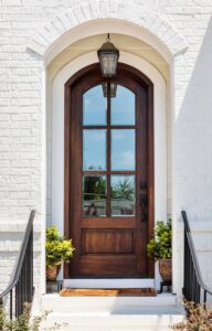 The exterior of a white brick home with an arched entryway. An arched wooden door with six windows is below a pendant light.