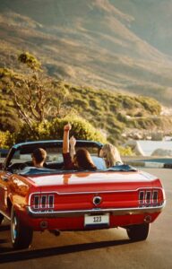 A bright red convertible driving down the road on a sunny day. Two passengers hold their hands in the air.
