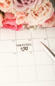 A white calendar with the text "wedding" written on the 18th of the month. A bouquet of pink roses lies on the calendar.