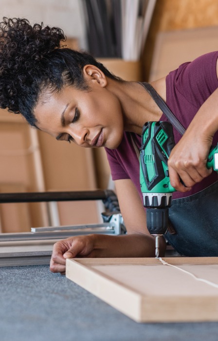 Woman with curly hair uses a cordless power drill to secure a wooden panel on a workbench in a woodworking shop.