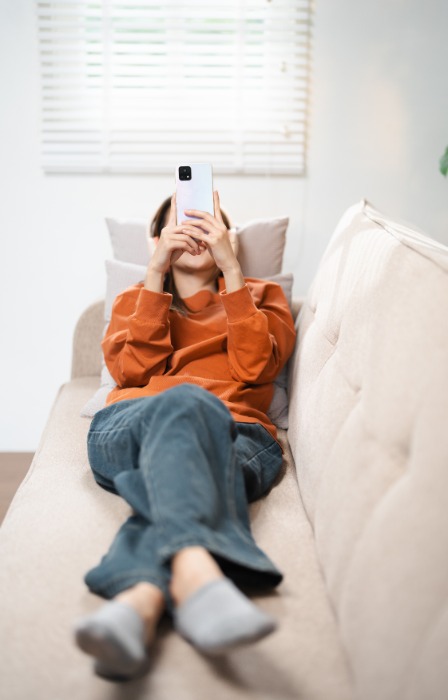 A woman wearing gray socks, sweatpants, and an orange shirt is lying on a white couch and looking at her phone.