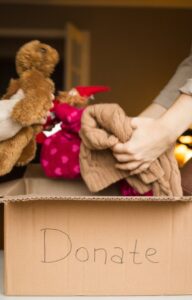 A young girl places a teddy bear into a donation box while an adult adds folded clothing beside her at home.
