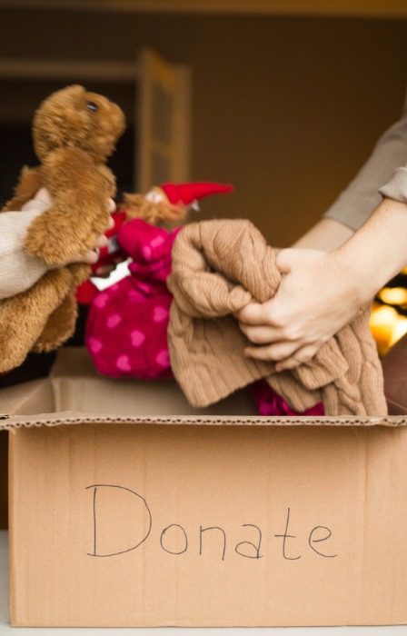 A young girl places a teddy bear into a donation box while an adult adds folded clothing beside her at home.